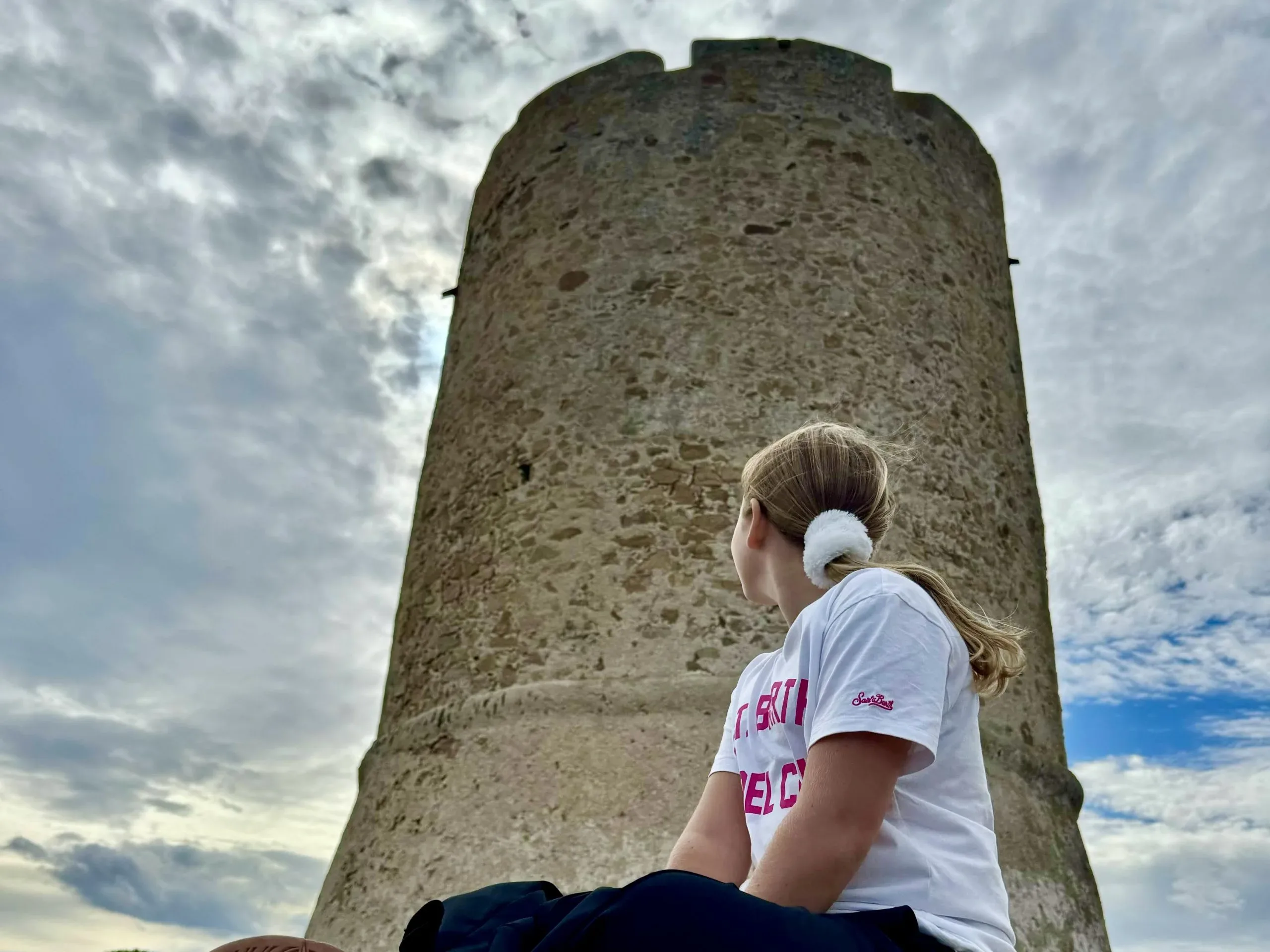 Girl looking at stone tower under cloudy sky.