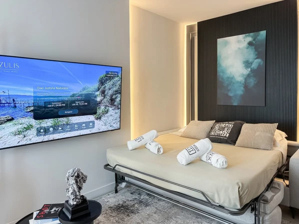 Camera da letto in stile suite d'albergo moderna con letto matrimoniale, asciugamani bianchi arrotolati, cuscini a righe, TV a parete con paesaggio costiero e opera d'arte con nuvole incorniciata su parete scura con effetto texture e illuminazione soffusa