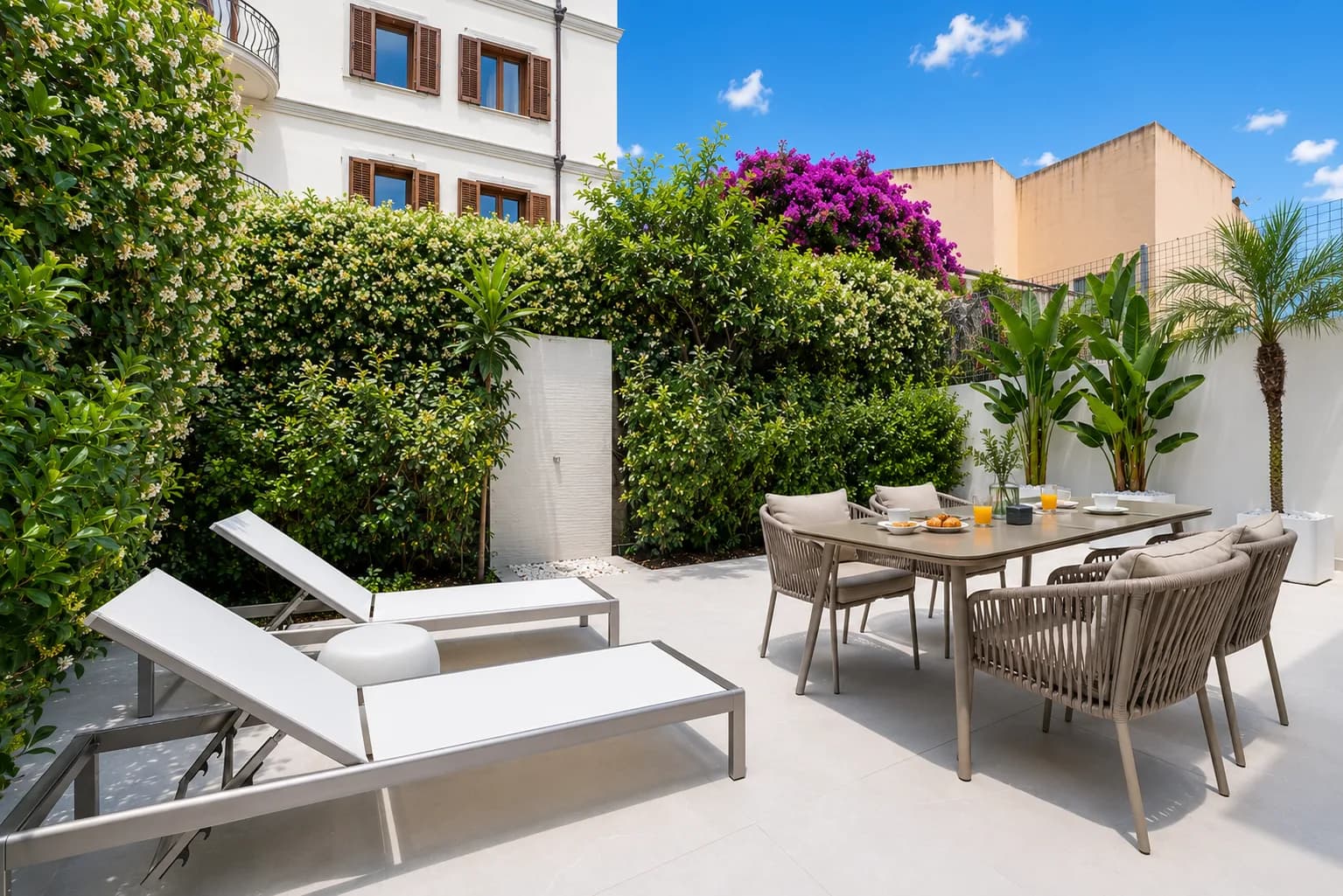 Terrazza giardino soleggiata con lettini bianchi, tavolo da pranzo apparecchiato per la colazione, rigogliosa edera rampicante e bouganvillea magenta, facciata bianca dell'edificio e palme sullo sfondo, Olbia, Sardegna