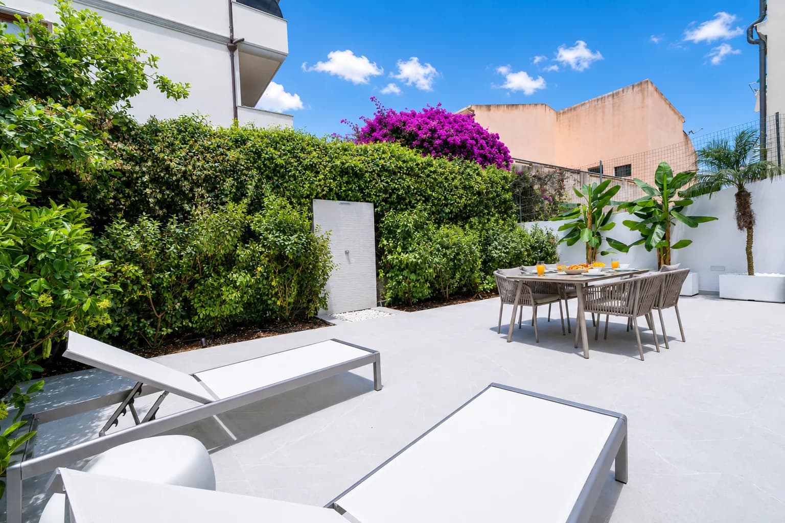 Terrazza giardino privata soleggiata con lettini bianchi, tavolo da pranzo, rigogliosa siepe verde, vivace bouganvillea magenta e architettura sarda contemporanea a Olbia