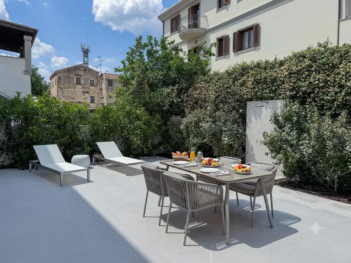 Terrazza di appartamento moderno con giardino a Olbia con tavolo da pranzo apparecchiato per la colazione, lettini, pareti coperte d'edera ed edifici d'epoca adiacenti sotto un cielo azzurro