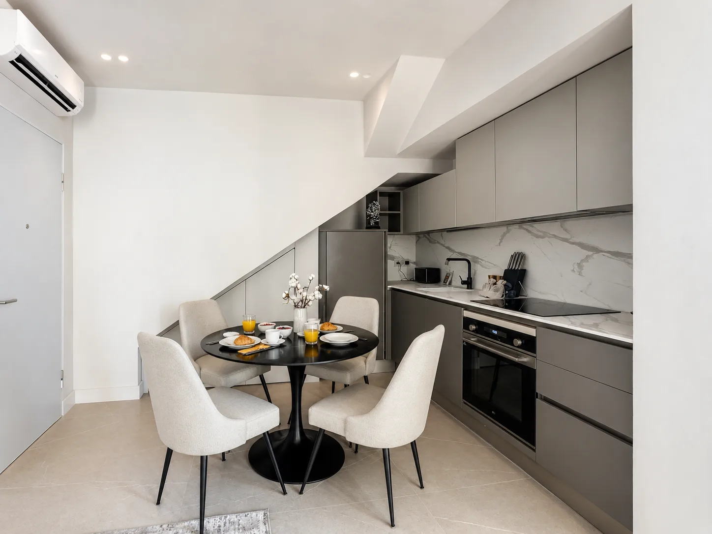 Zona cucina-pranzo moderna con mobili grigi, schienale in marmo, tavolo rotondo nero con sedie color crema e posate apparecchiate nell'appartamento AZULIS DelleTerme Suites, Olbia