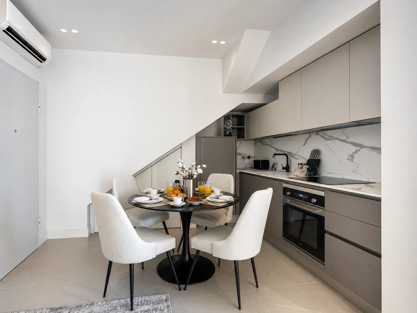 Zona cucina-pranzo moderna con mobili color crema e tortora, schienale in marmo, tavolo da pranzo rotondo nero con due sedie color crema e colazione con succo d'arancia e frutta