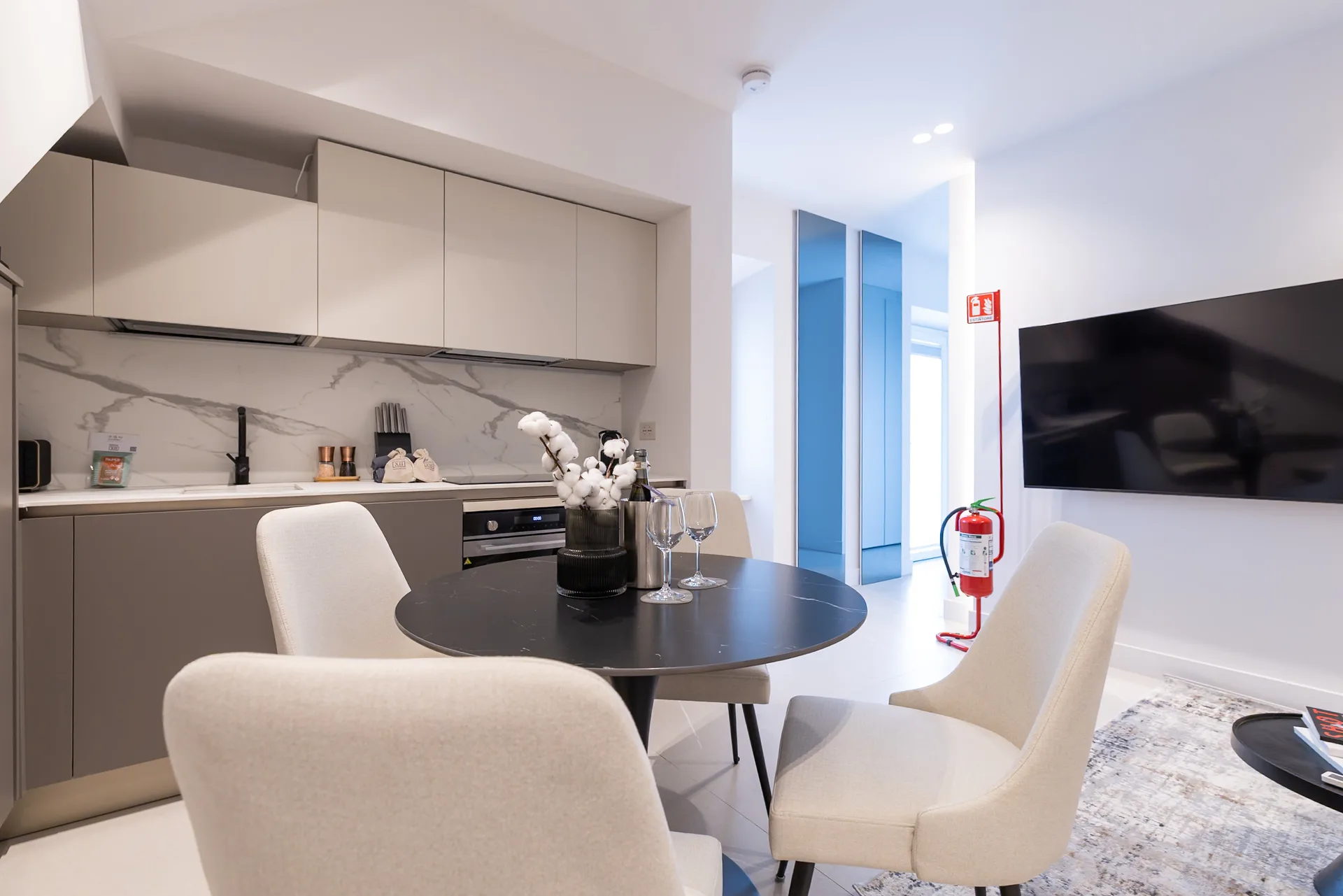 Interno di appartamento moderno open-space con cucina integrata, zona pranzo con tavolo rotondo nero e sedie color crema, e area salotto con TV a parete e porte blu che conducono al giardino