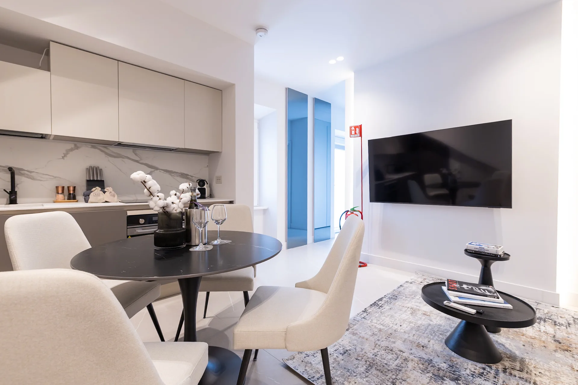 Zona soggiorno open-space moderna con sedie da pranzo color crema attorno a un tavolo rotondo nero, cucina con mobili bianchi, televisione a parete e porte a vetri blu che conducono a uno spazio esterno
