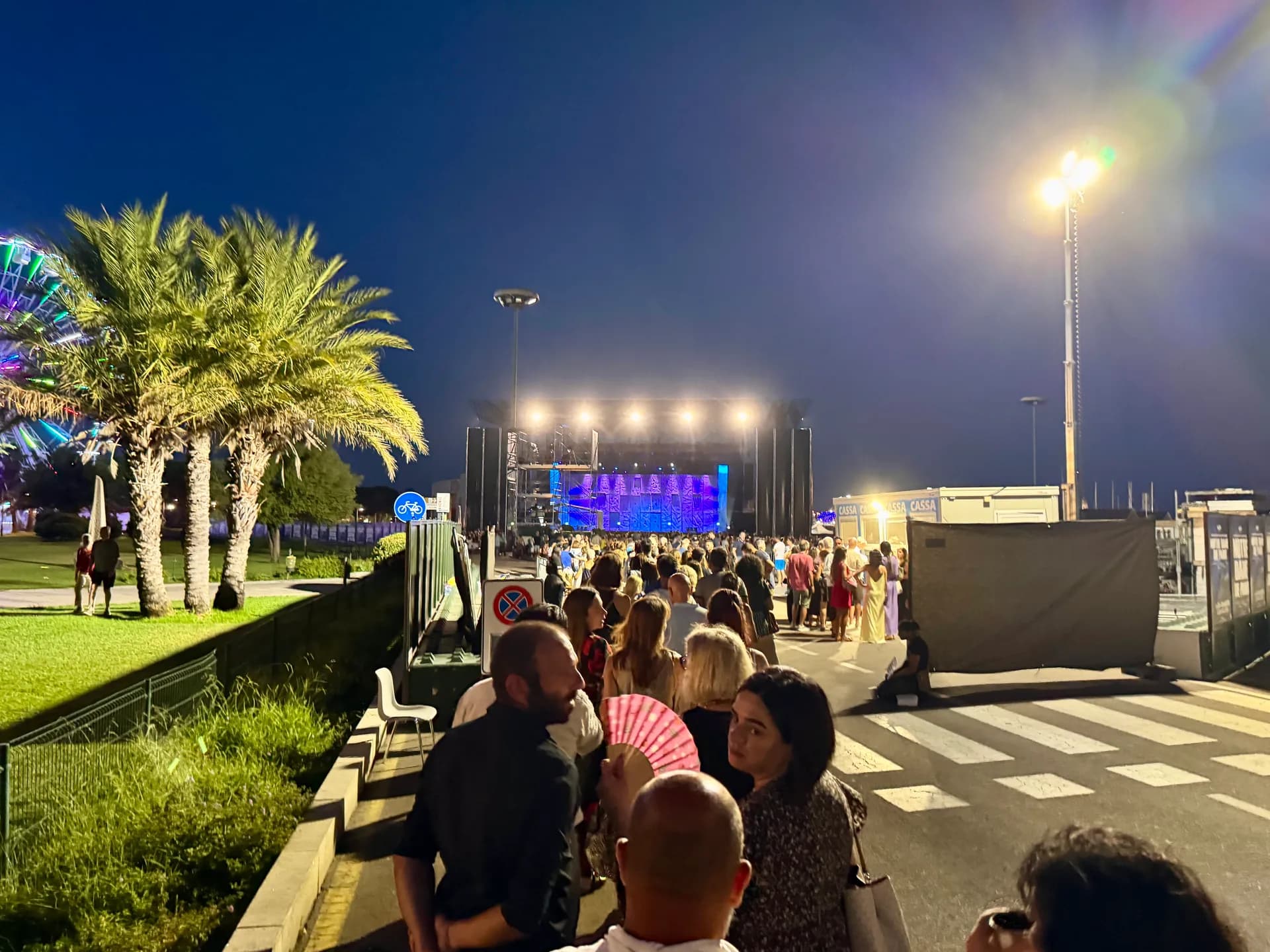 Crowds gather at Molo Brin waterfront in Olbia at dusk, with an illuminated stage displaying purple and blue lights, palm trees lining the left side, and street lighting overhead