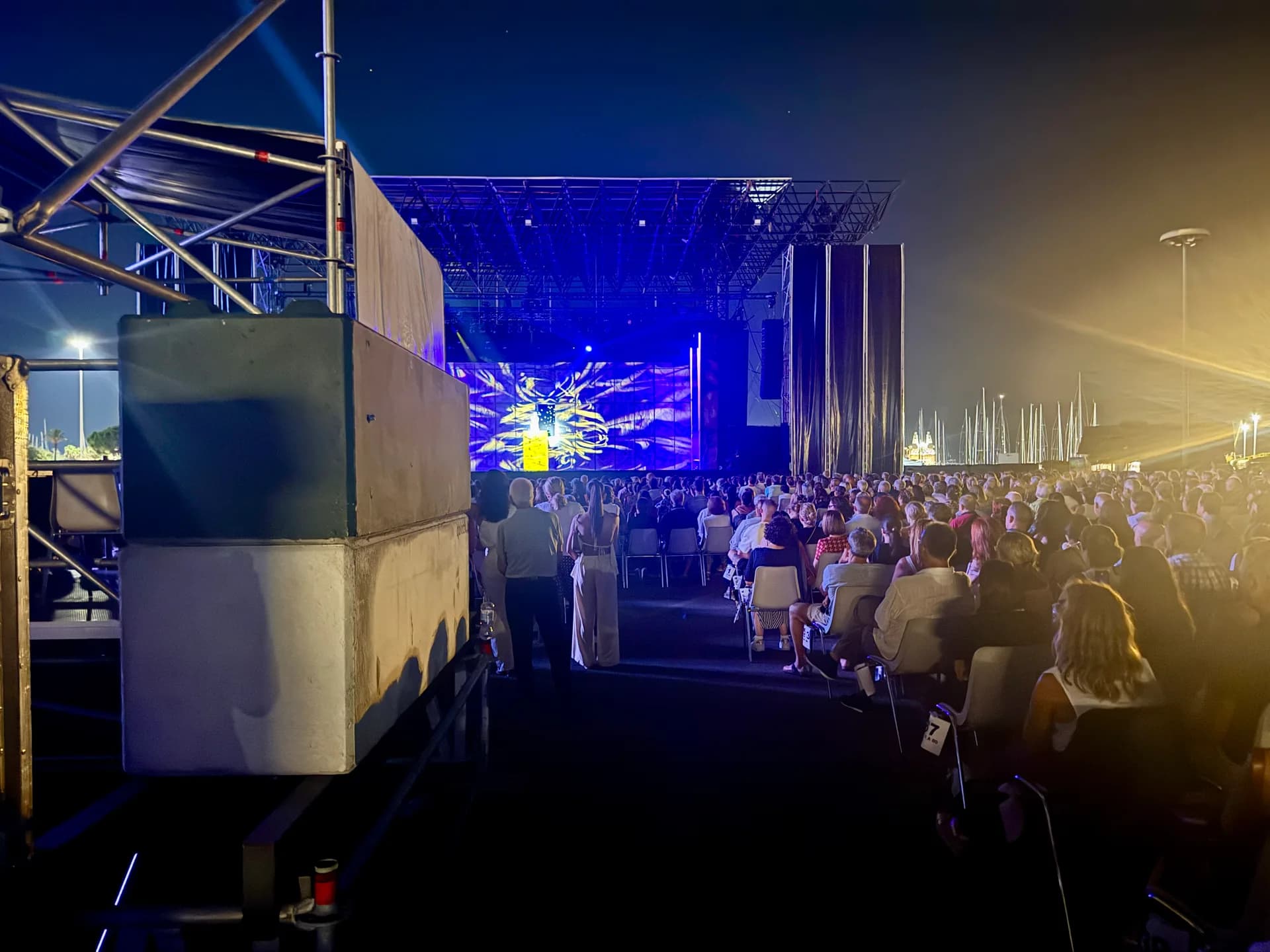 Nighttime outdoor concert at Molo Brin, Olbia, with large crowd facing illuminated stage displaying blue light projection and cathedral imagery