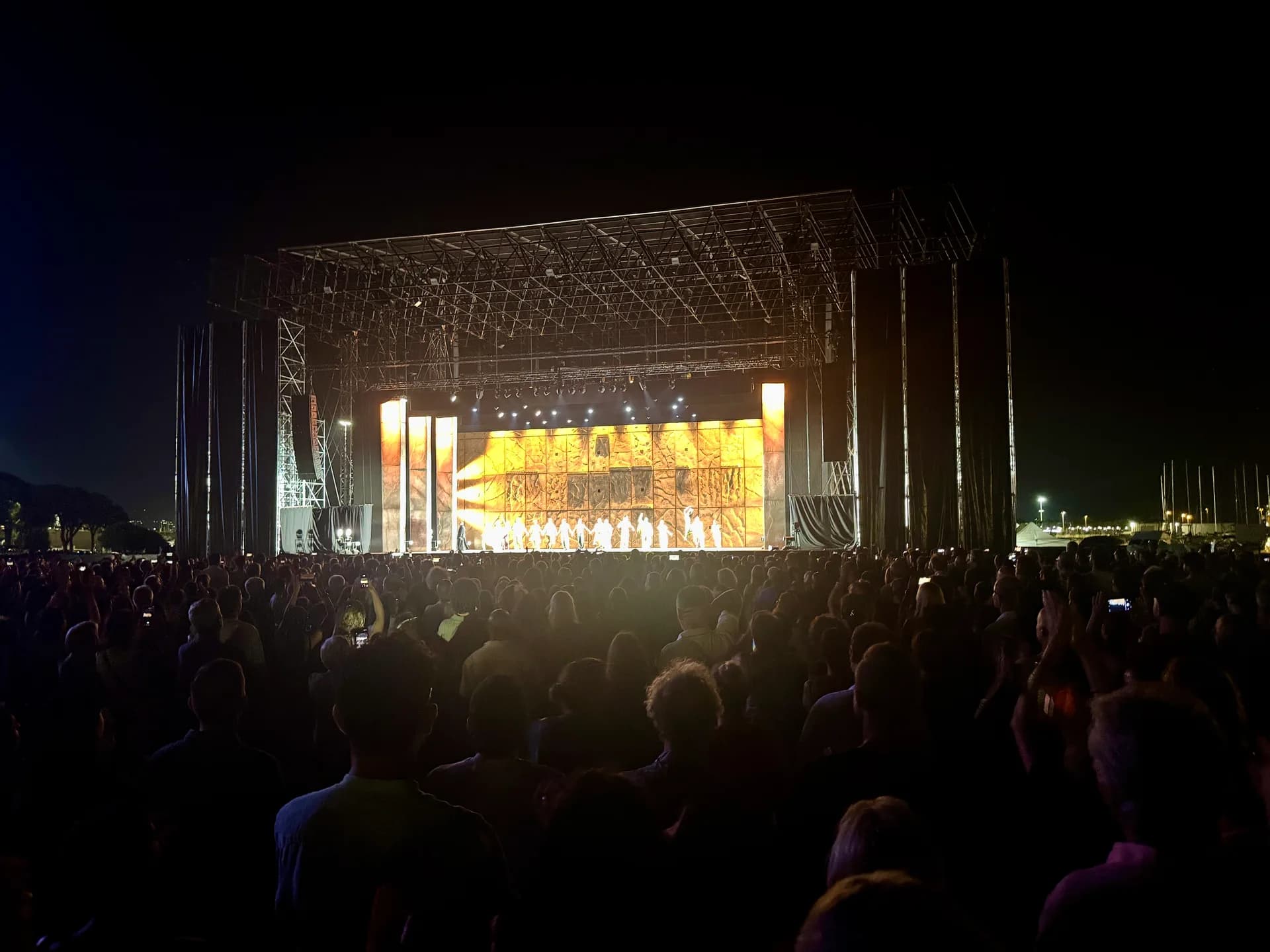 Outdoor concert stage at night with large illuminated screen showing Notre-Dame cathedral imagery, crowd of spectators in foreground at Molo Brin, Olbia