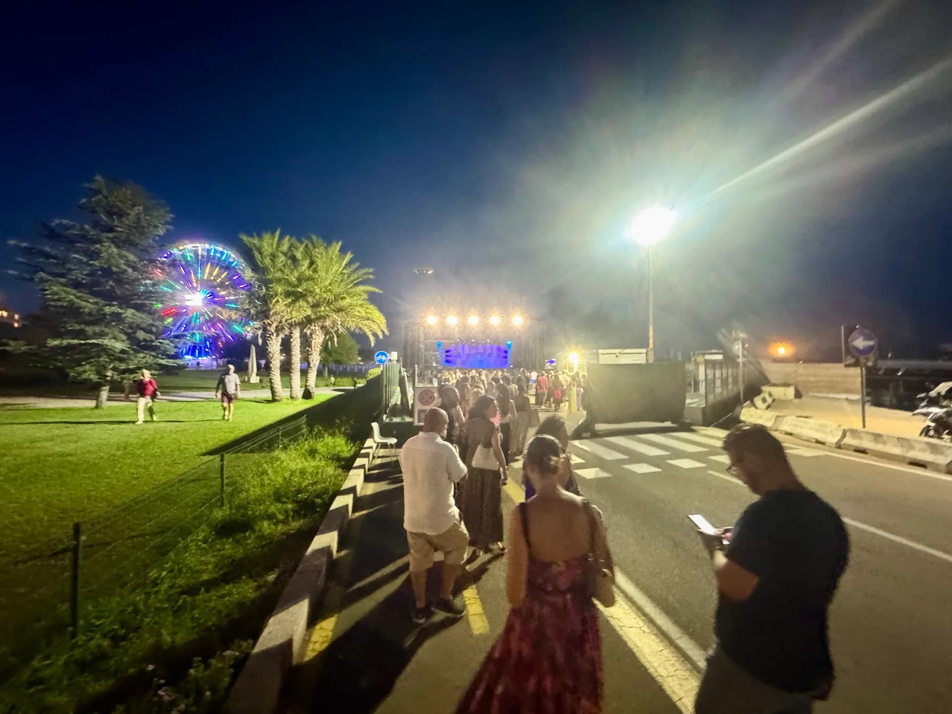 Evening crowd walking along a waterfront promenade in Olbia with illuminated Ferris wheel, palm trees, and stage lights visible in the background