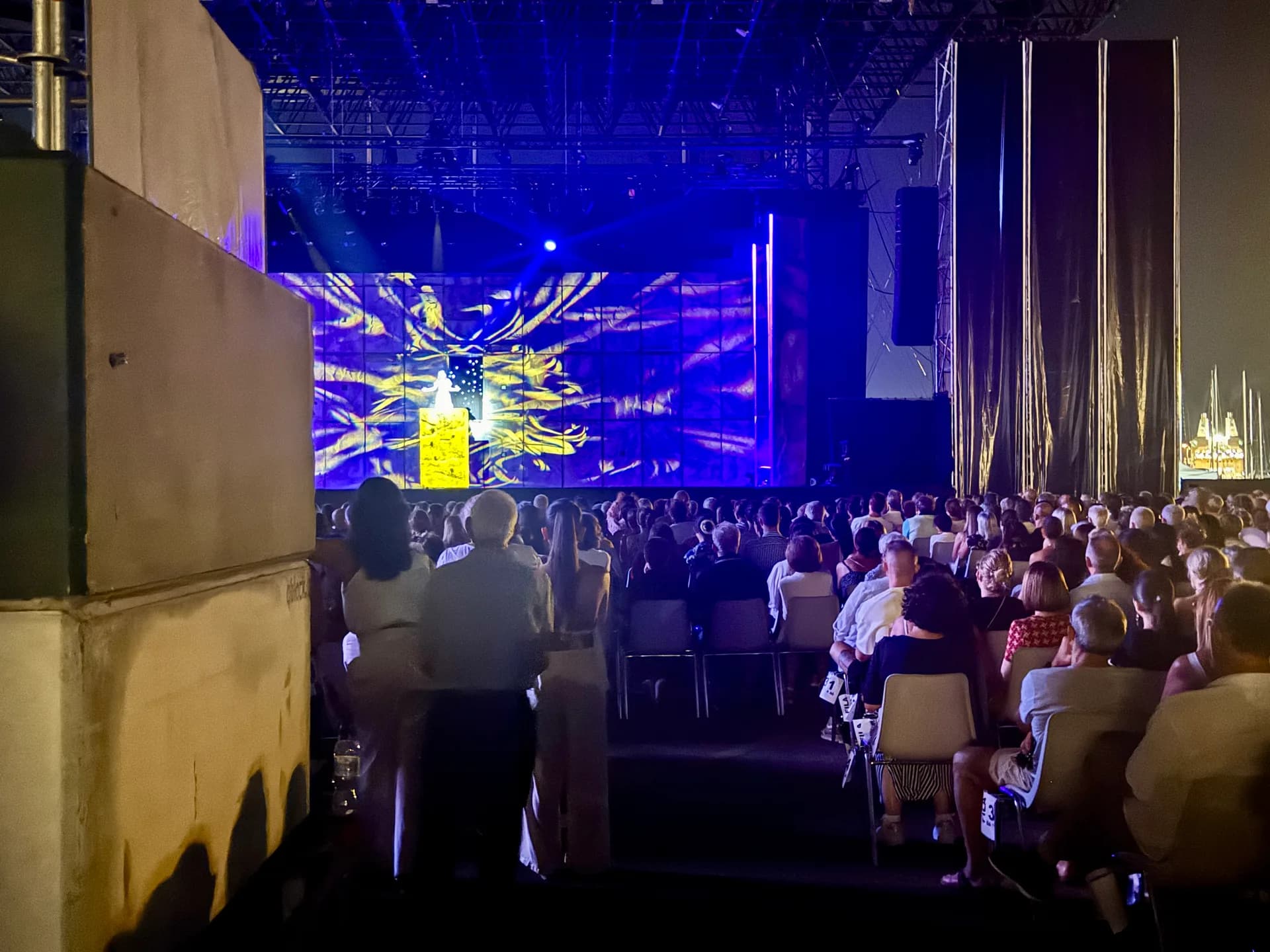 Outdoor concert venue at Molo Brin, Olbia, with audience seated facing a large illuminated screen displaying Notre-Dame de Paris in yellow and blue lighting against a night sky