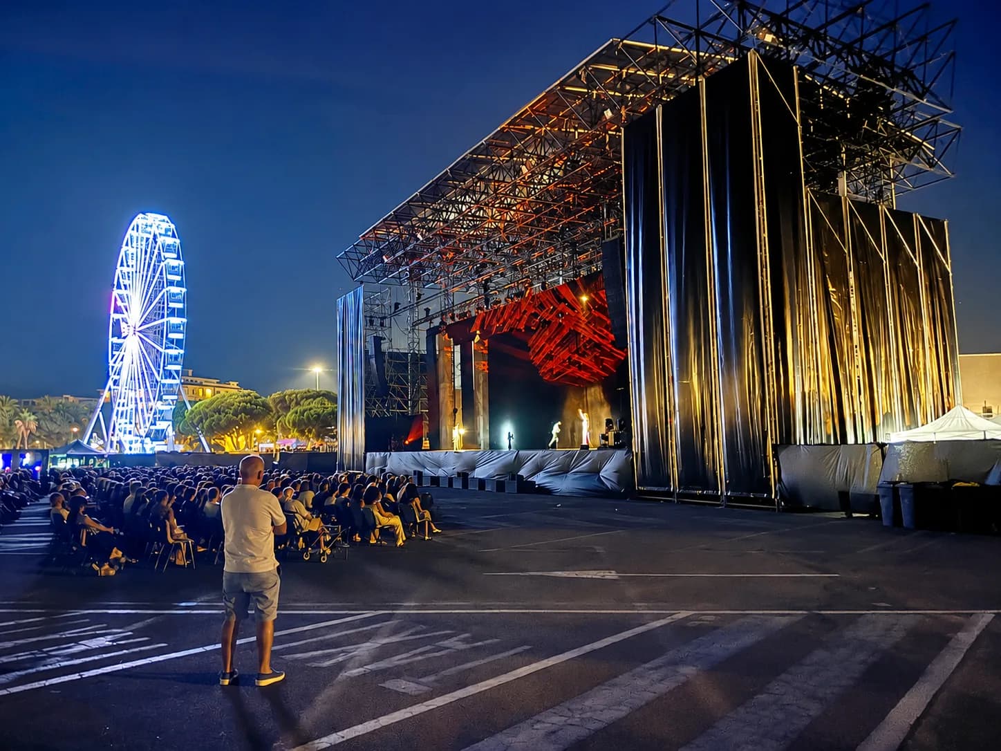 Evening event at Molo Brin waterfront in Olbia with illuminated Ferris wheel, large outdoor stage with red lighting, and seated audience under blue twilight sky