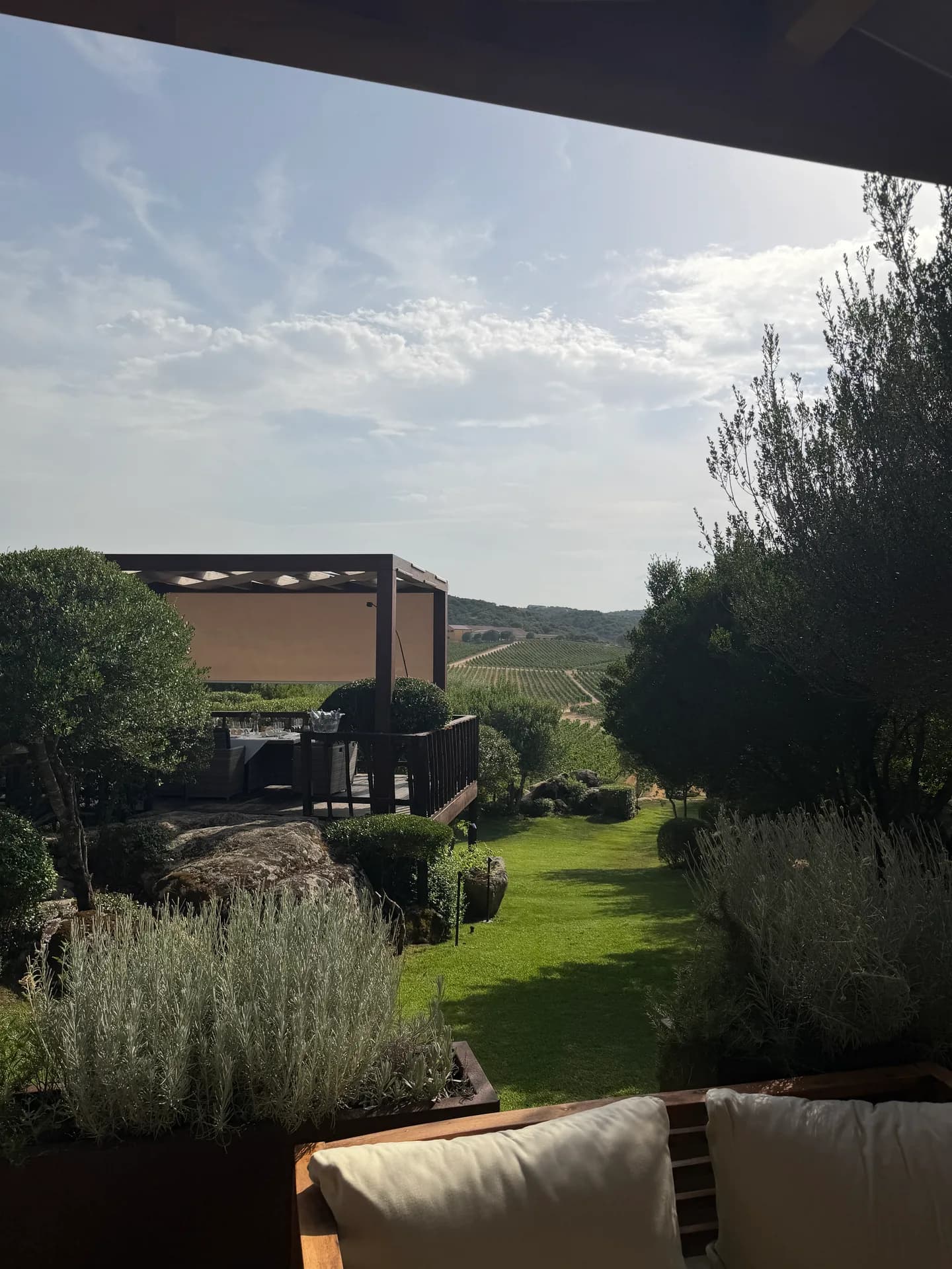 Modern pergola with retractable shade overlooking manicured gardens and Sardinian vineyards under a partly cloudy sky