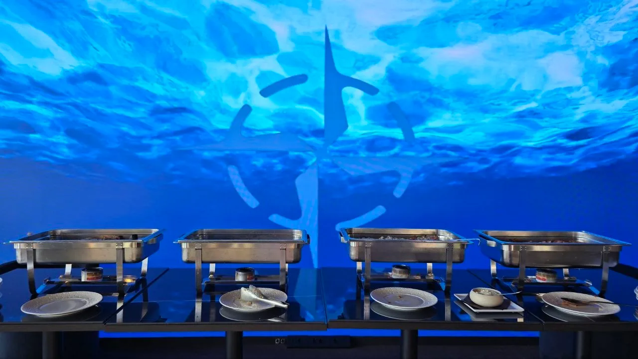 Buffet chafing dishes and plates lined up against a striking blue underwater-themed backdrop at Nord Est restaurant in Olbia, Sardinia.
