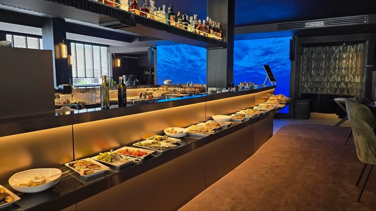 Curved buffet counter at Nord Est Olbia lined with dishes of antipasti, backlit under warm lighting with a blue underwater-themed screen behind the bar.
