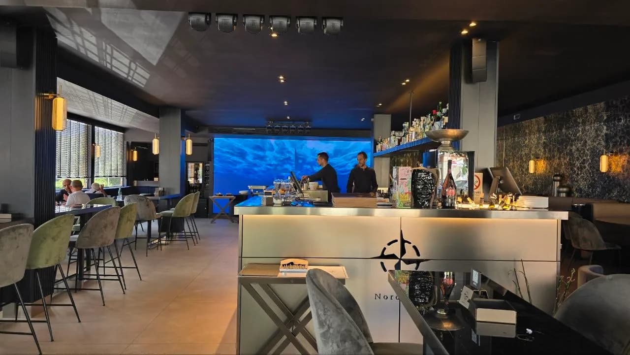 Sleek modern interior of Nord Est restaurant in Olbia, with a stainless steel bar, velvet stools, and staff working beneath a large screen displaying a blue underwater scene.