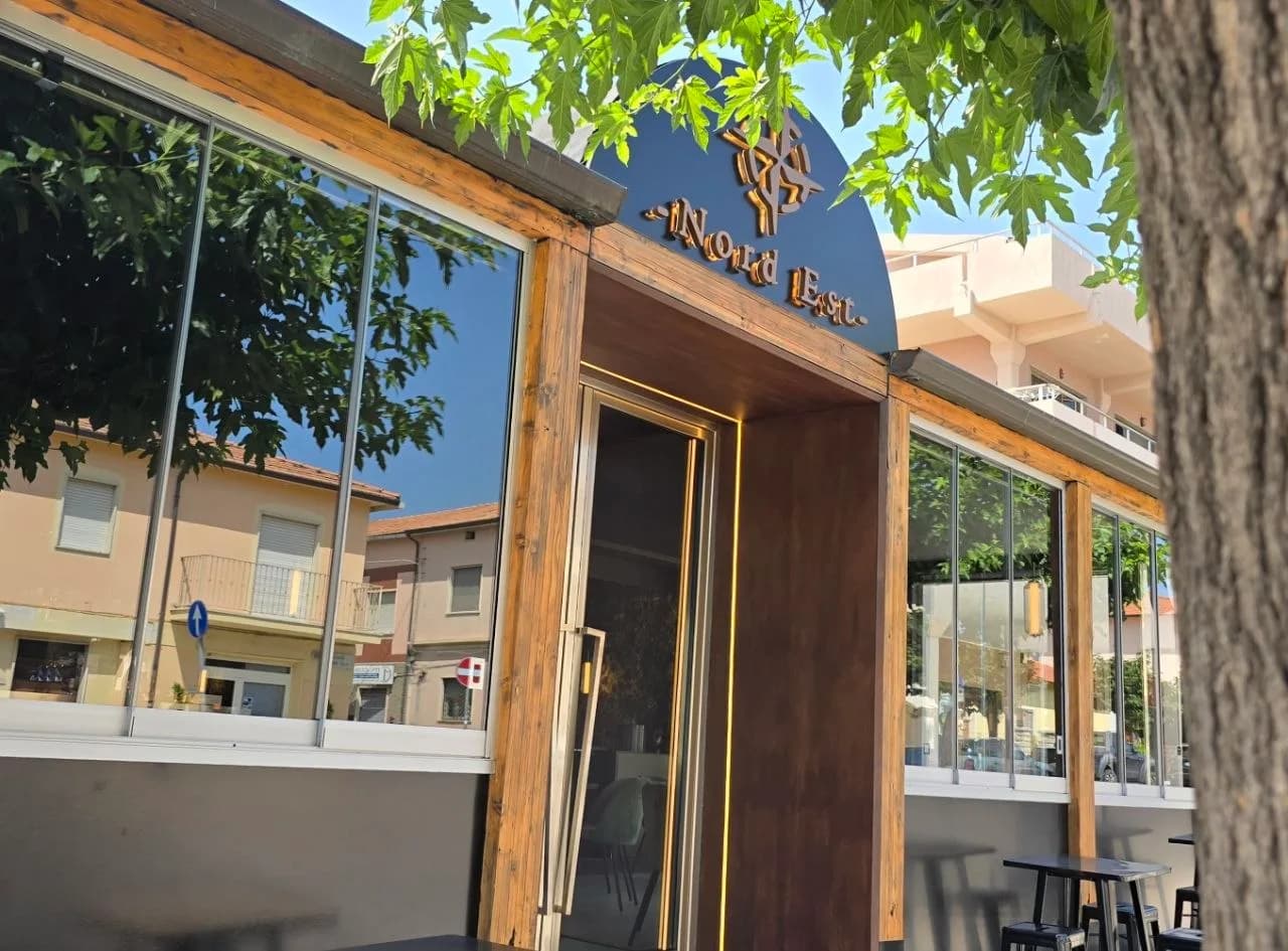 Wooden-framed glass front of Nord Est restaurant in Olbia, with an arched sign above the entrance and street buildings reflected in the windows.