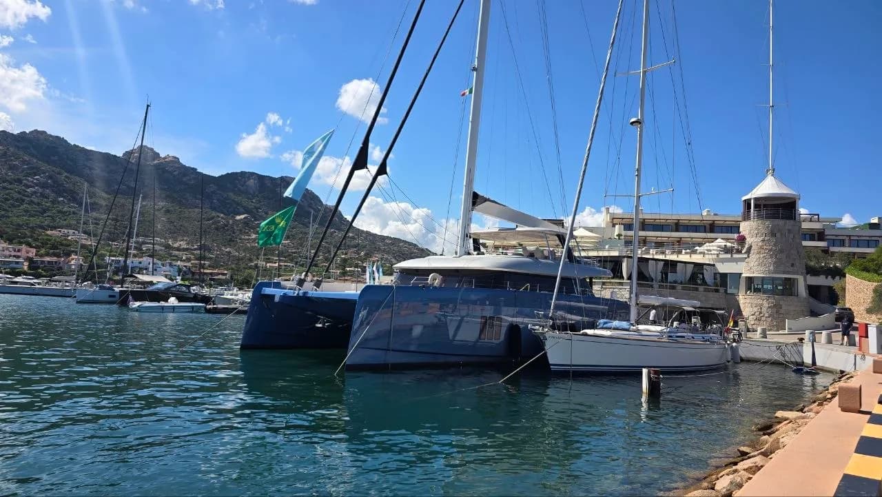 Sailing yachts moored in Porto Cervo marina, with a stone lighthouse tower, hillside villas and Sardinian mountains in the background.