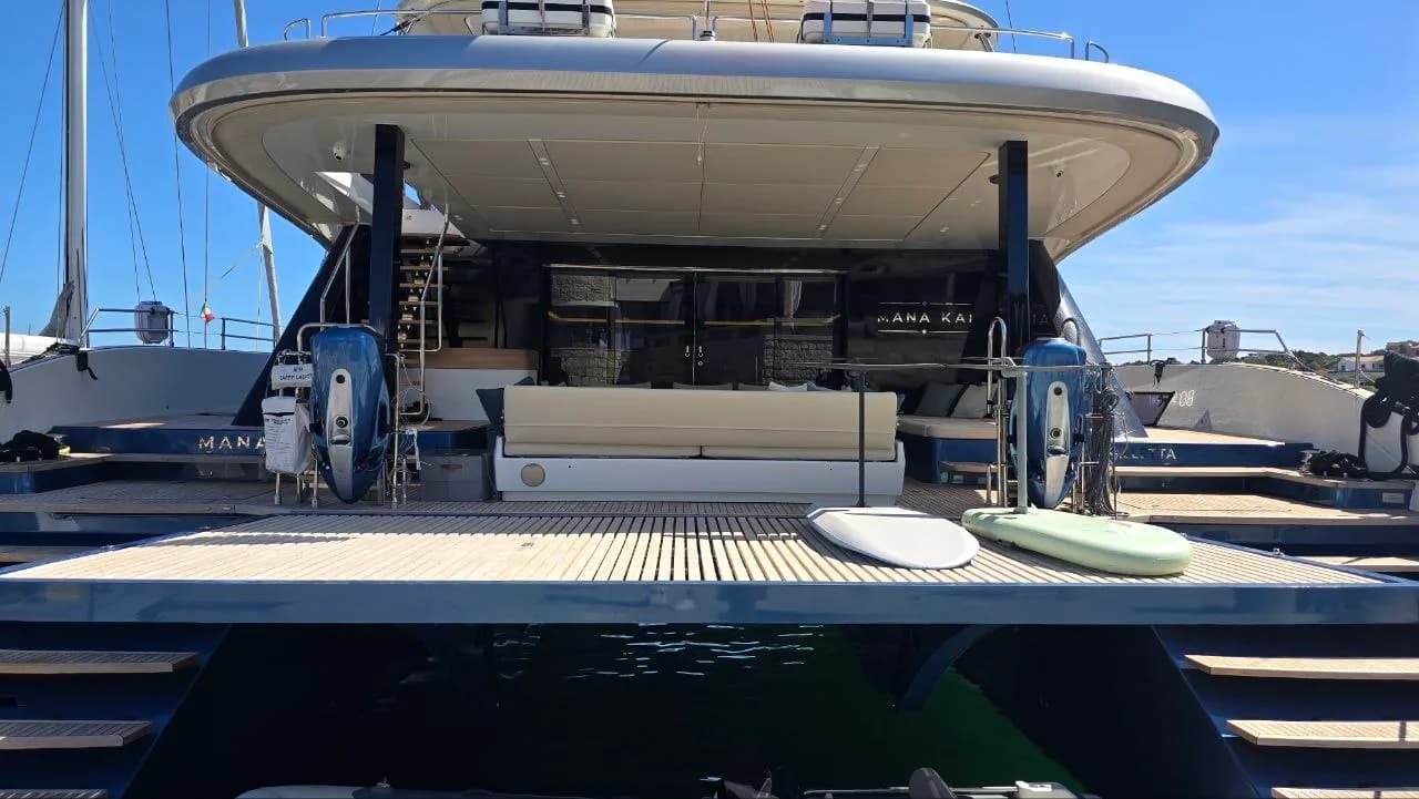 Rear deck of a luxury motor yacht moored in Porto Cervo marina, with teak flooring, cream seating, jet skis and paddleboards under a shaded canopy.