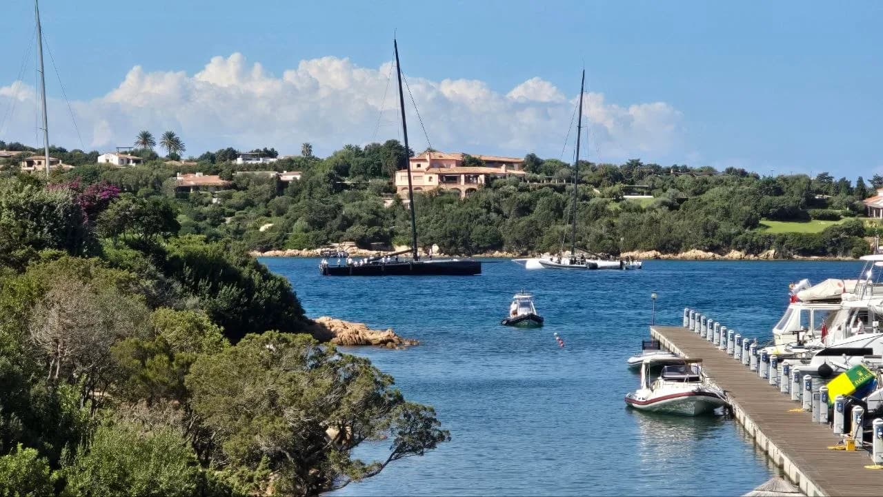 Tall-masted maxi yachts moored in the bay off Porto Cervo, with a marina jetty, small motorboats and wooded coastline in the background