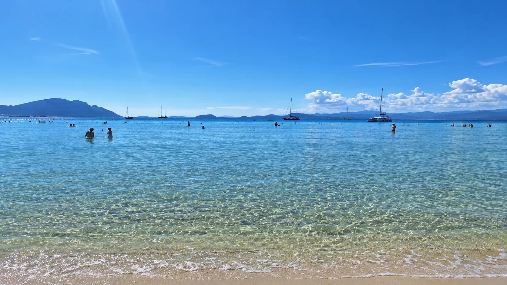Swimmers in the clear turquoise water of Terza Spiaggia, Golfo Aranci, with sailboats moored offshore and hills lining the horizon under a blue sky.