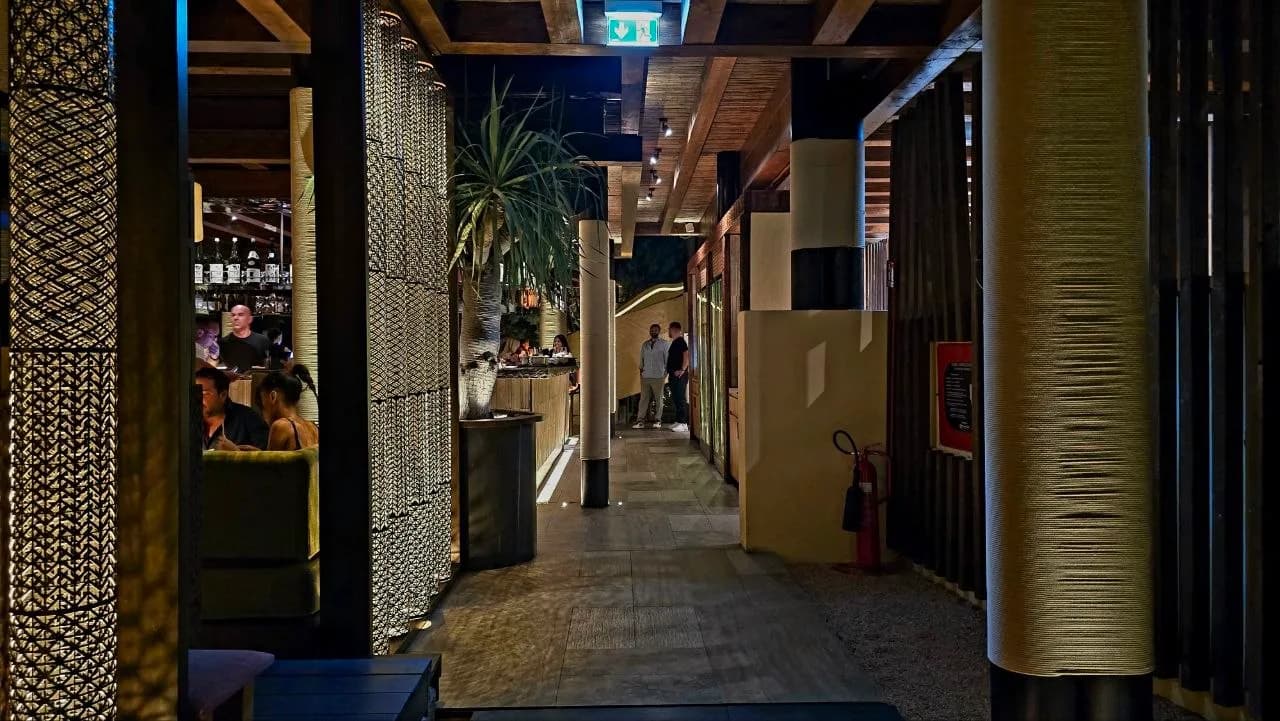Dimly lit interior walkway at Zuma Porto Cervo at night, with woven textured columns, wooden beams, a potted palm and guests seated at the bar and standing along the corridor.