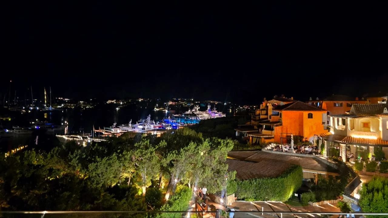 Night view over Porto Cervo marina with illuminated yachts, terracotta buildings and diners on a terrace above the harbour