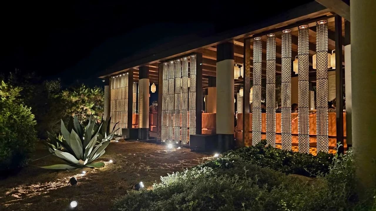 Night view of Zuma Porto Cervo's terrace, with chainmail-curtained columns, warm lantern lighting and an agave plant in the garden foreground.