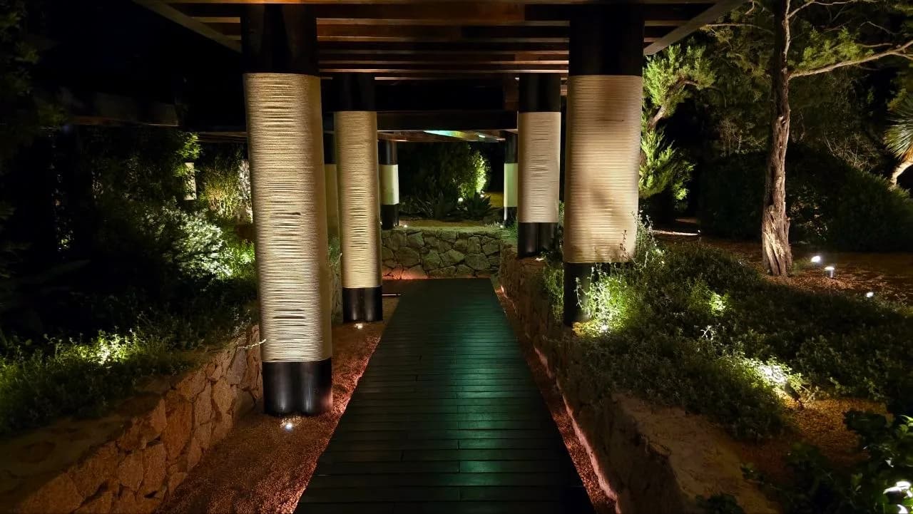 Wooden decked walkway at night, lined with rope-wrapped columns under a pergola and softly lit Mediterranean gardens, leading towards Zuma Porto Cervo.