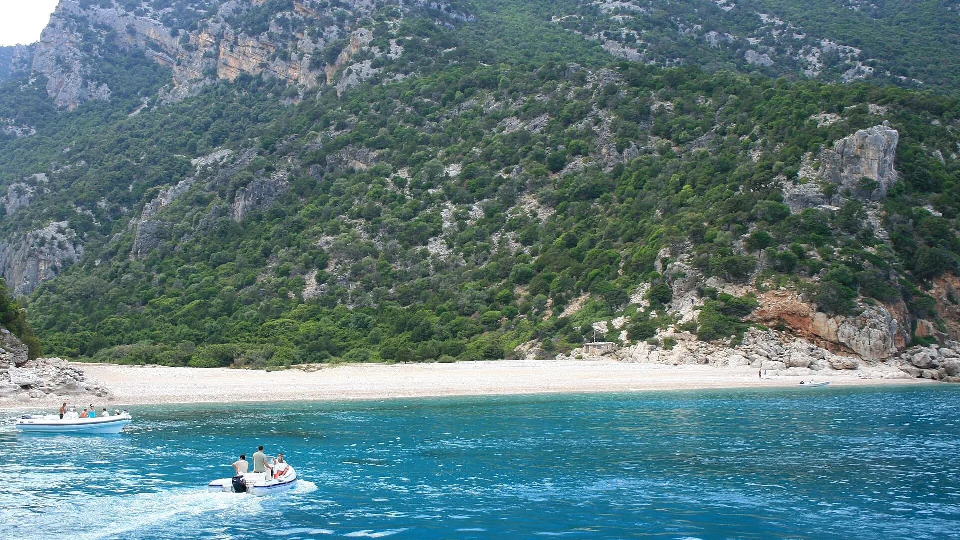 Two inflatable boats crossing turquoise water towards the long pebble beach of Cala Sisine under wooded limestone slopes