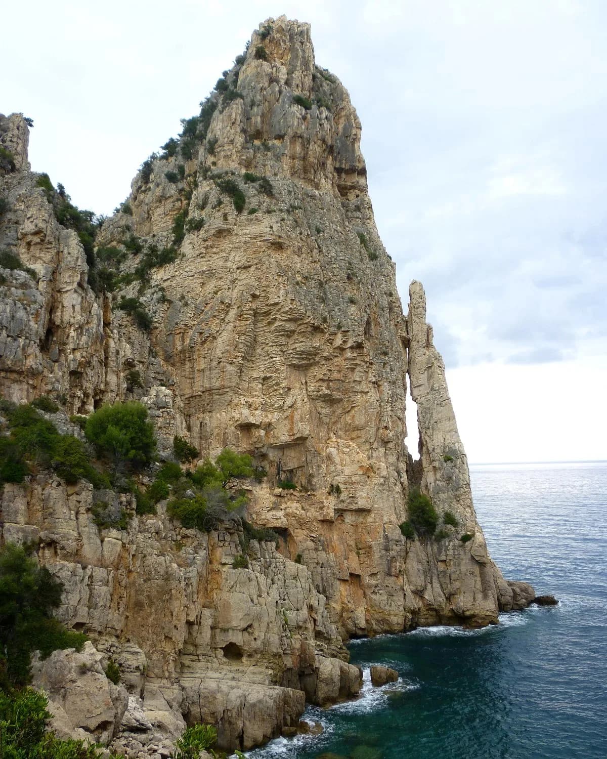 Pedra Longa, a tall pale limestone pinnacle rising straight from the sea on the Baunei coast, under a cloudy sky