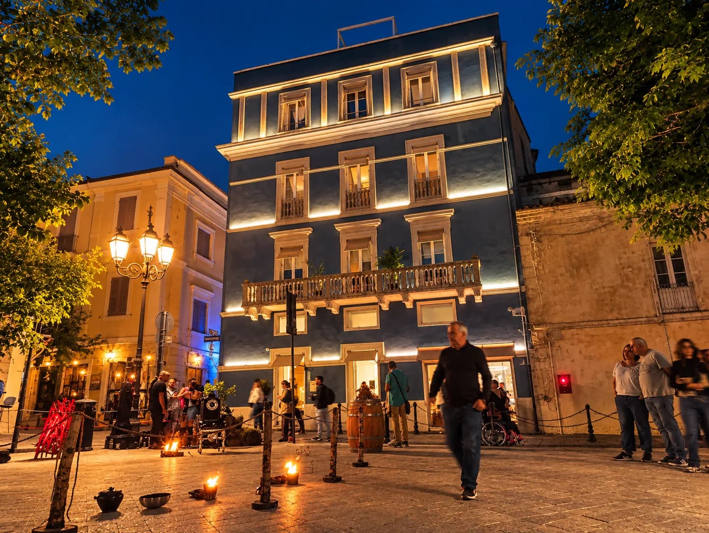 Een groot houten wijnvat met het opschrift 'Benvenuto Vermentino Olbia' staat op het oude stadsplein van Olbia bij schemering, terwijl festivalgangers wijn drinken onder bomen versierd met lichtsnoeren.