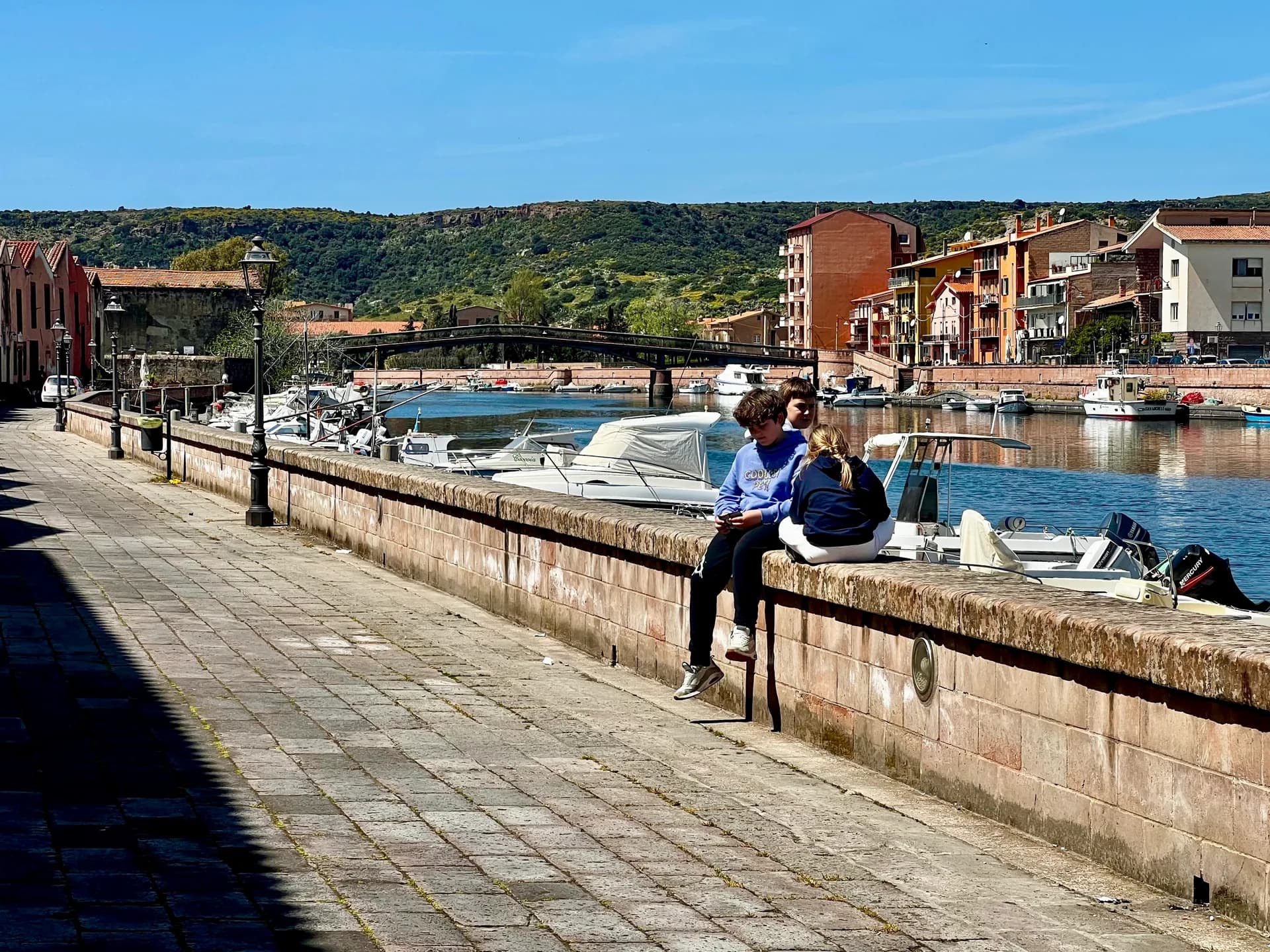 Children sit on a stone riverside wall overlooking moored boats on the Temo river in Bosa, with colourful buildings and hills in the background.