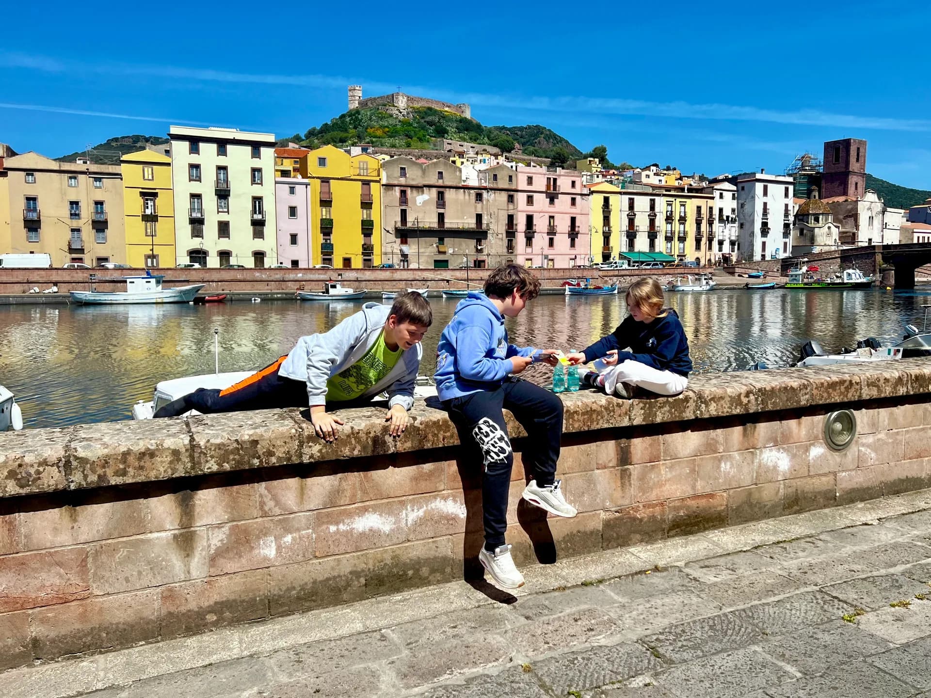 Children sitting on a riverside wall in Bosa, Sardinia, with colourful houses, moored boats and the hilltop Malaspina Castle in the background.