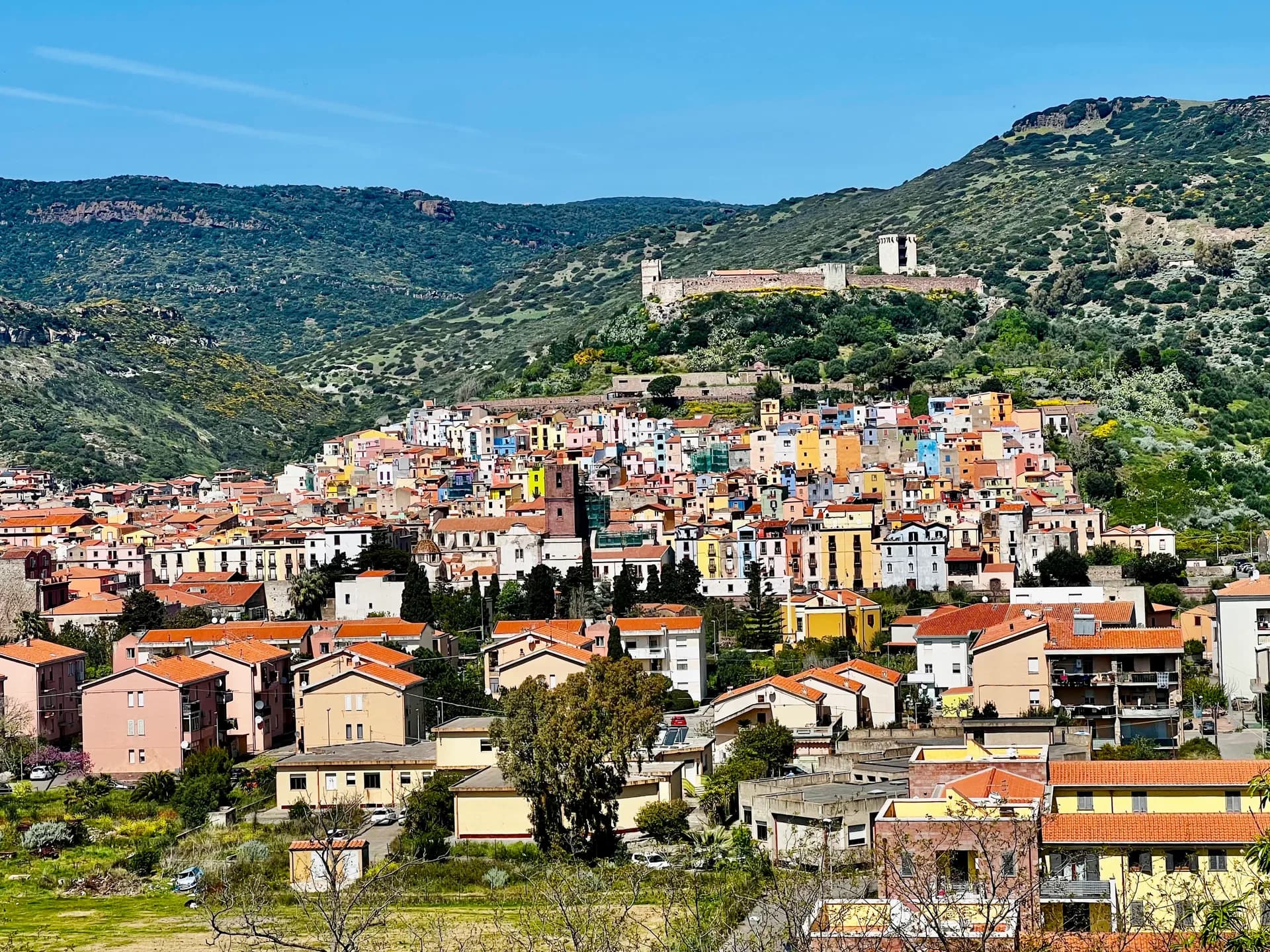 View over Bosa's pastel-coloured houses with terracotta roofs climbing the hillside, overlooked by the ruined medieval Malaspina Castle and green hills beyond.