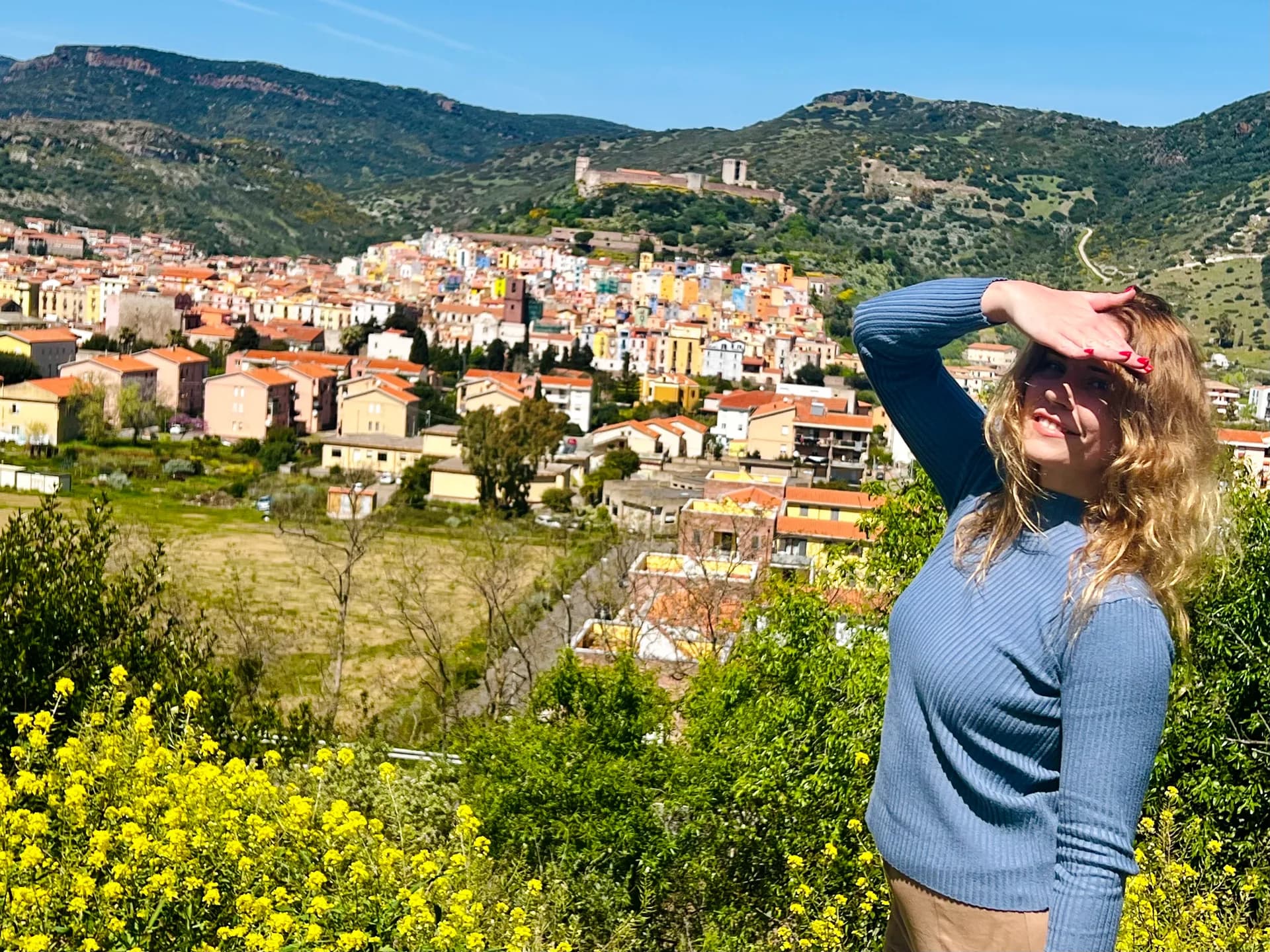A woman shields her eyes from the sun amid yellow wildflowers, overlooking Bosa's colourful hillside houses, the Malaspina Castle and green mountains beyond.