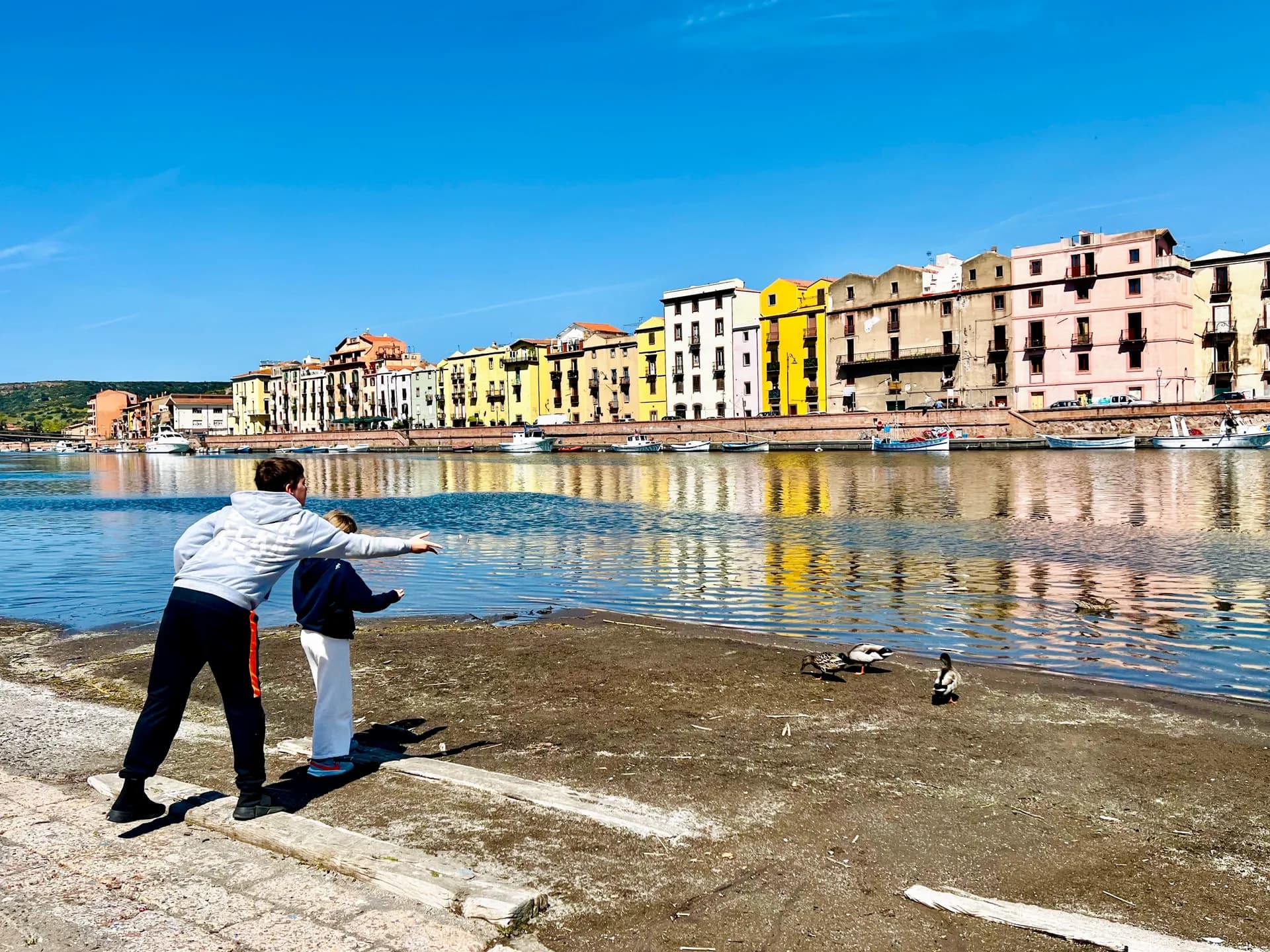 Two boys point towards ducks by the Temo river in Bosa, Sardinia, with colourful houses, moored boats and their reflections lining the far bank.