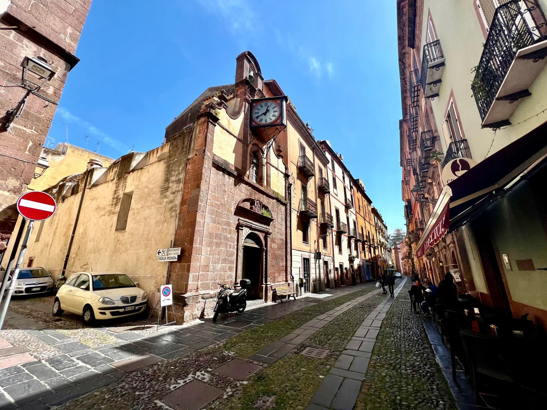 Narrow cobbled street in Bosa's old town, lined with pastel-coloured buildings, a stone church with a projecting clock, parked cars and scooter, and café tables further down the lane.