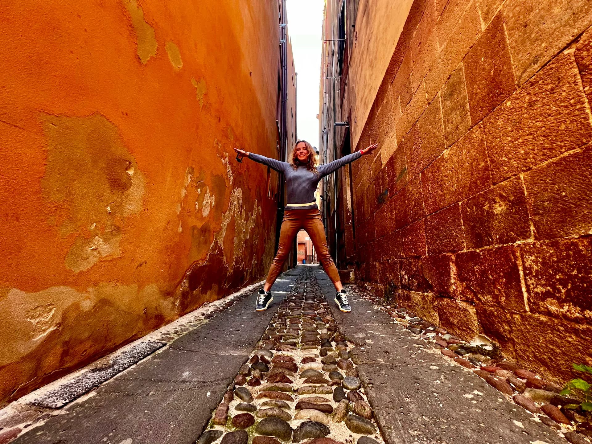 Woman jumping with arms and legs outstretched in a narrow cobbled alley between weathered orange and terracotta walls in Bosa, Sardinia