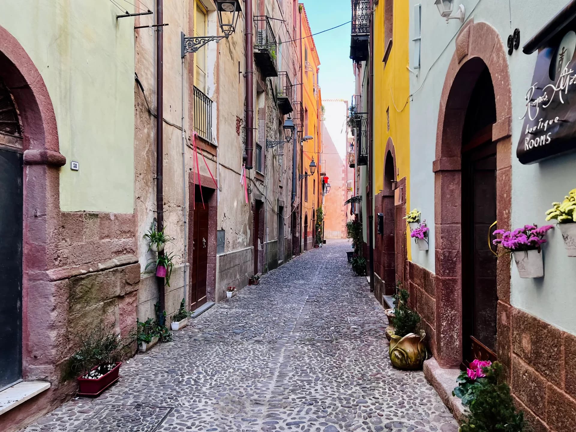 Narrow cobbled street in Bosa, Sardinia, lined with pastel-coloured houses, stone archways and potted flowers.