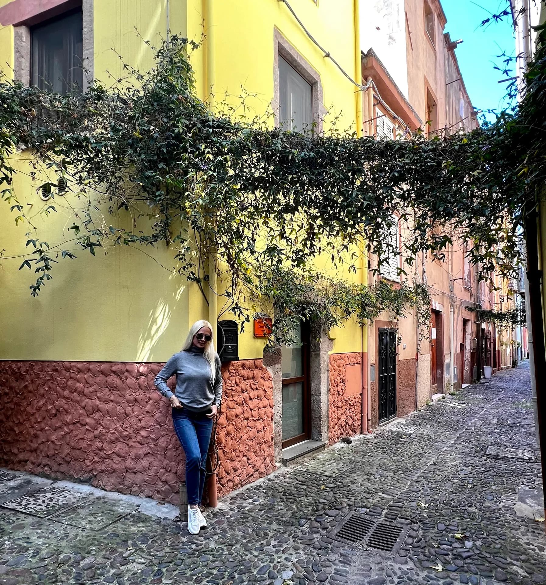 A woman leans against a yellow and terracotta house on a cobbled street in Bosa, Sardinia, with trailing green vines above and colourful buildings lining the lane.