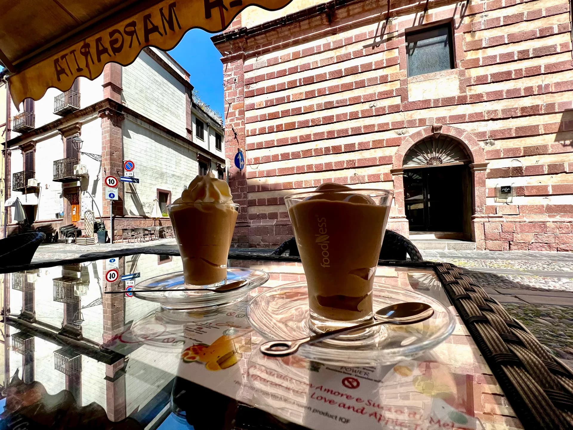 Two iced coffees on a glass café table in a cobbled street in Bosa, Sardinia, with the town's striped brick buildings behind.