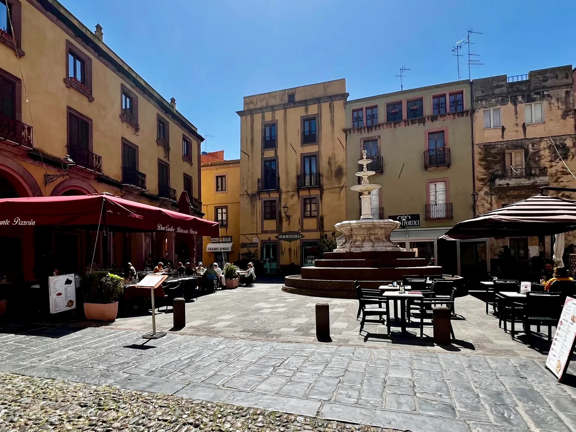 Sunny piazza in Bosa with a tiered stone fountain, pastel-coloured buildings, and pavement cafe tables beneath red umbrellas.