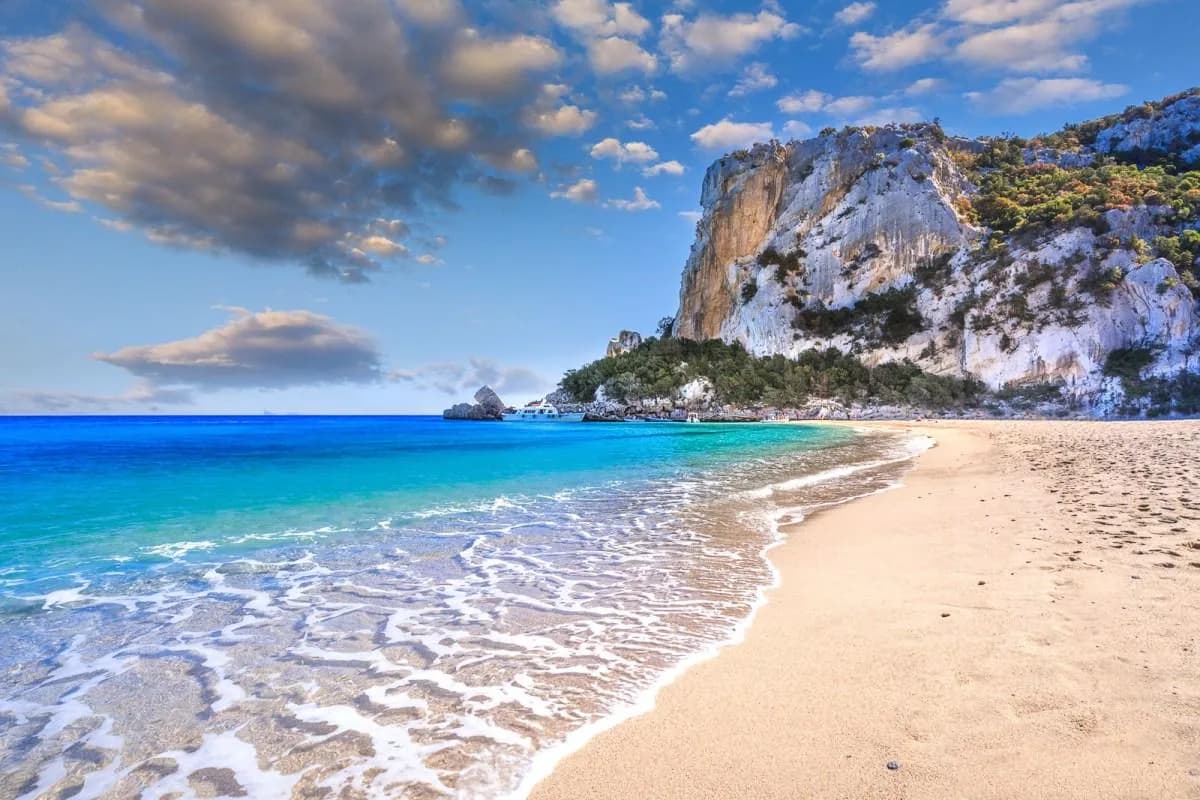 Turquoise sea lapping onto the sandy shore of Cala Luna, with pale limestone cliffs and green scrub rising behind the Gulf of Orosei beach.