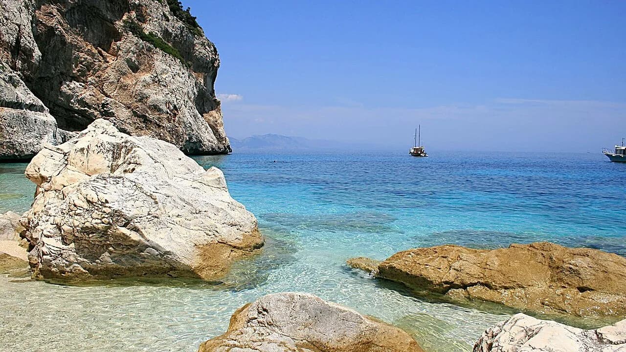 Pale limestone boulders in clear turquoise shallows at Cala Mariolu, Sardinia, with two boats on the horizon