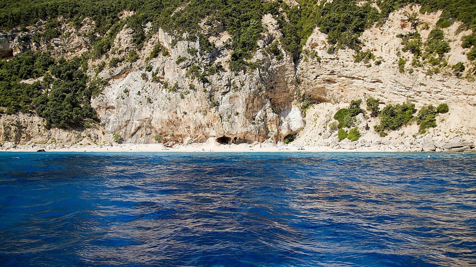 Cala dei Gabbiani on the Baunei coast seen from the sea: a white beach with a cave under scrub-covered limestone cliffs