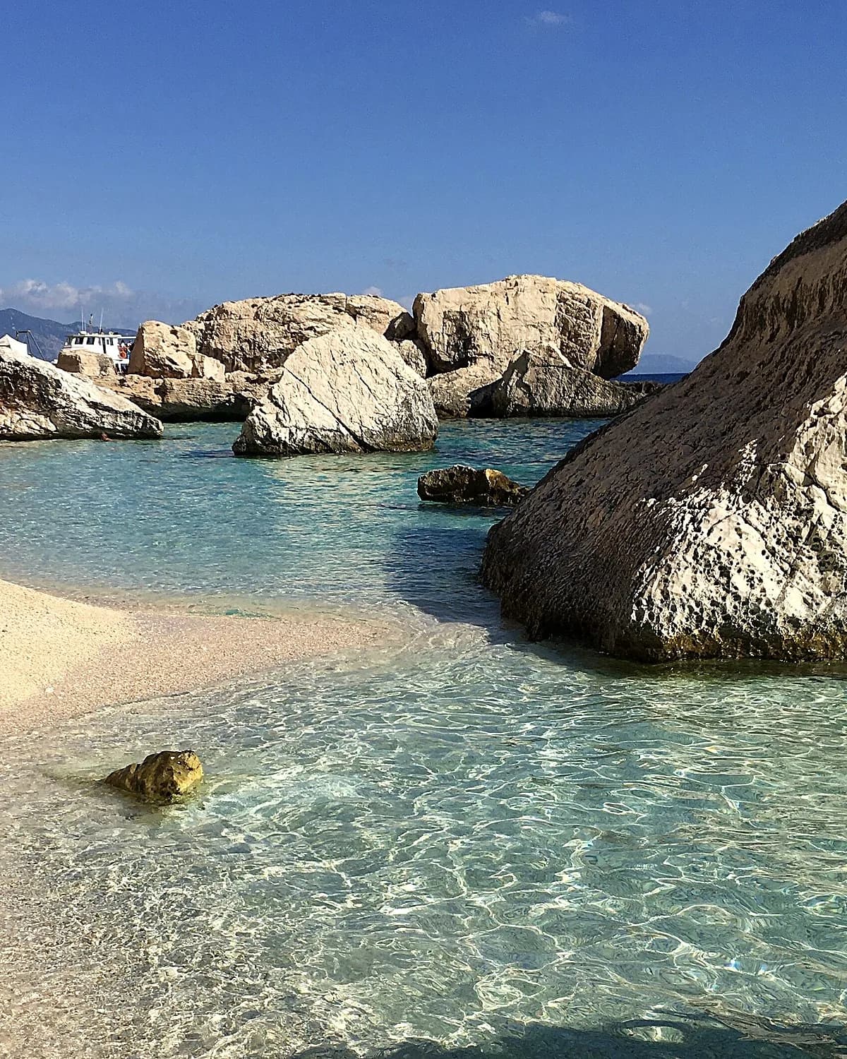 Large pale boulders and clear shallow water over a pebble shore at Cala Mariolu, Sardinia, with a tour boat behind