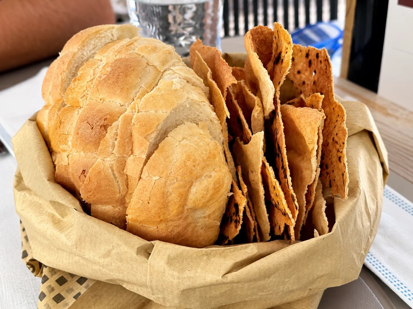 Sac en papier posé sur une table de restaurant, contenant une miche de pain blanc croustillante tranchée à côté de feuilles dressées de pain plat fin, croustillant et boursouflé.