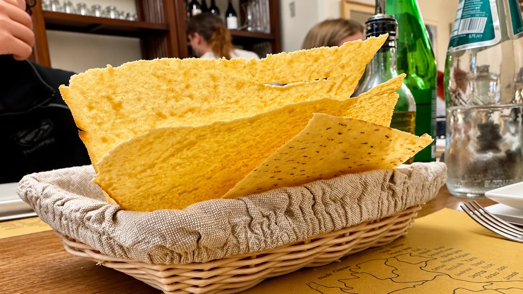 Feuilles de pain plat dorées, fines et croustillantes dans une corbeille en osier tapissée de lin sur une table de restaurant, avec des bouteilles, une fourchette et des convives flous à l'arrière-plan.