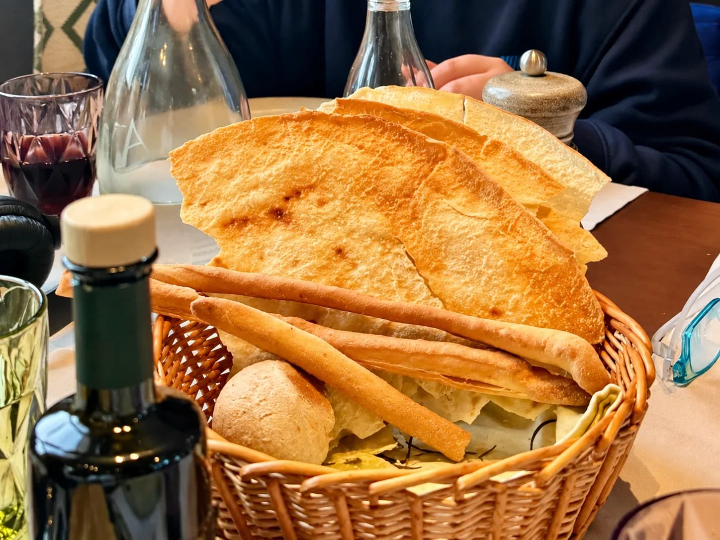 Corbeille en osier de feuilles de pain plat fines et croustillantes, de gressins et d'un petit pain sur une table de restaurant, avec une bouteille d'huile d'olive, une carafe d'eau et un verre de vin rouge.