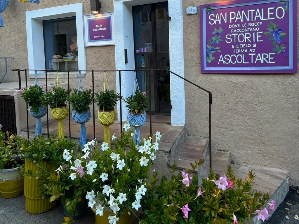 Shopfront in San Pantaleo with a purple painted sign, macramé hanging plant pots, flowering mandevilla in planters, and steps leading to an open doorway.